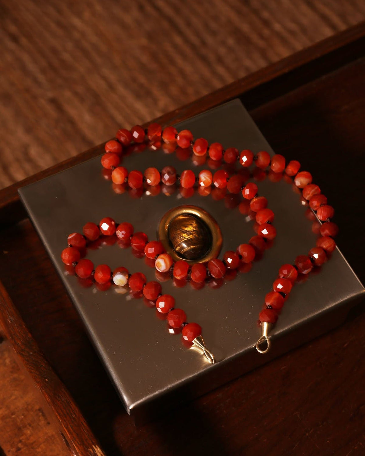 A beaded necklace made of faceted red beads with small gold-toned spacers and clasps, arranged on a dark metallic square tray over a wooden surface.